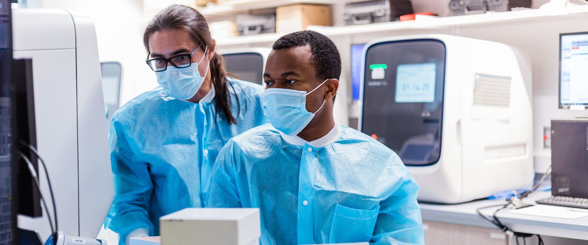 two people wearing masks look at a large device in a lab, with additional lab equipment behind them