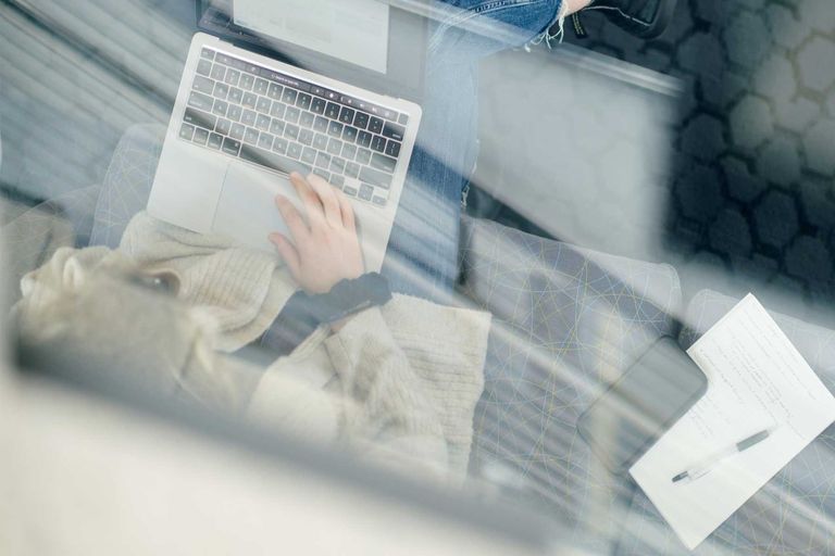 A view from above a person working on their laptop. They are sitting on a couch and have a black scrunchie on their wrist as they work on a grey and black laptop.