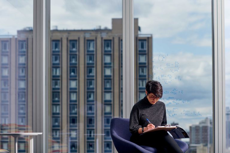 A person with short dark hair and glasses writes on a clipboard. They are sitting in front of a large window overlooking the BU East campus.