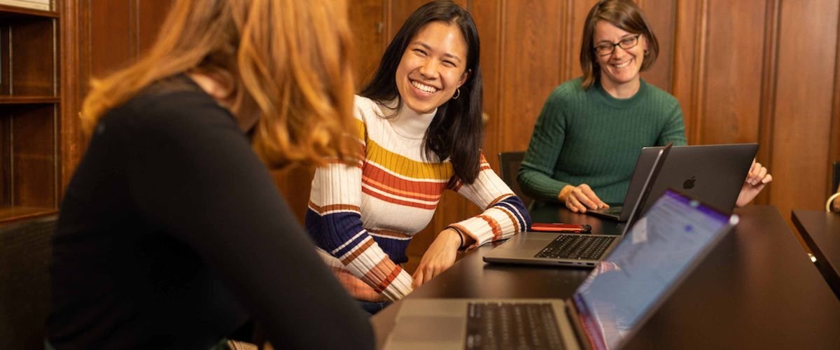 Three people sit around laptops in a wood paneled room. They are smiling and in conversation.