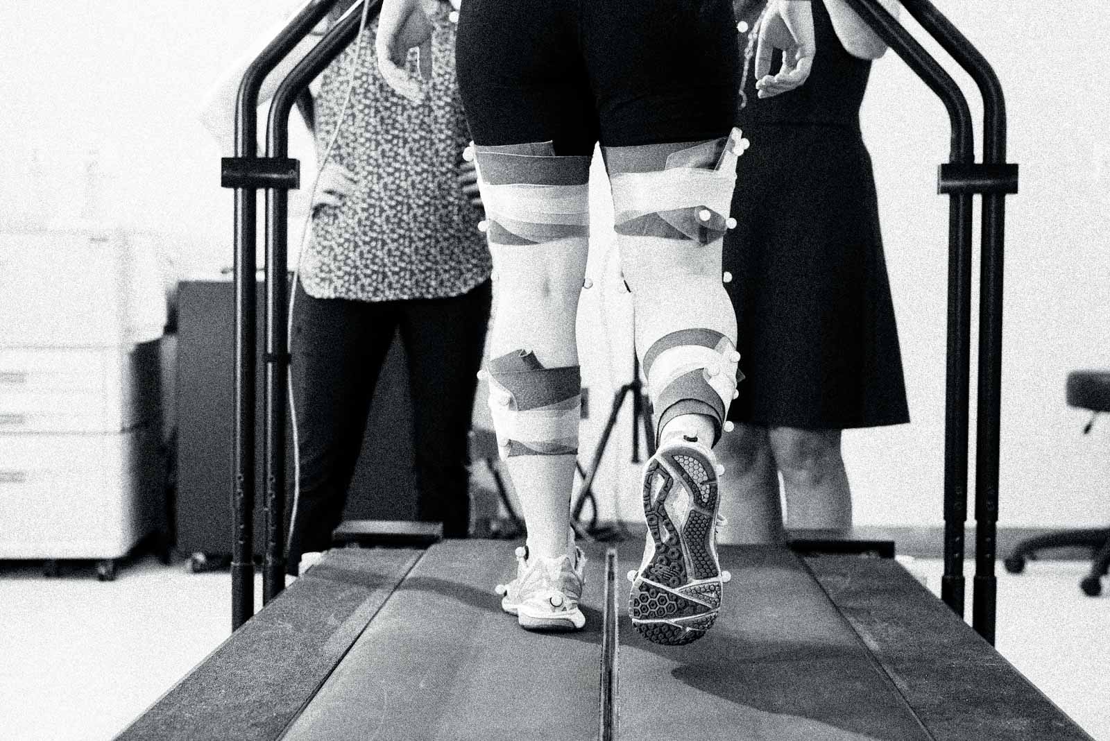 Back view of the legs of an individual walking on a treadmill, with the lower bodies of researchers visible in the background