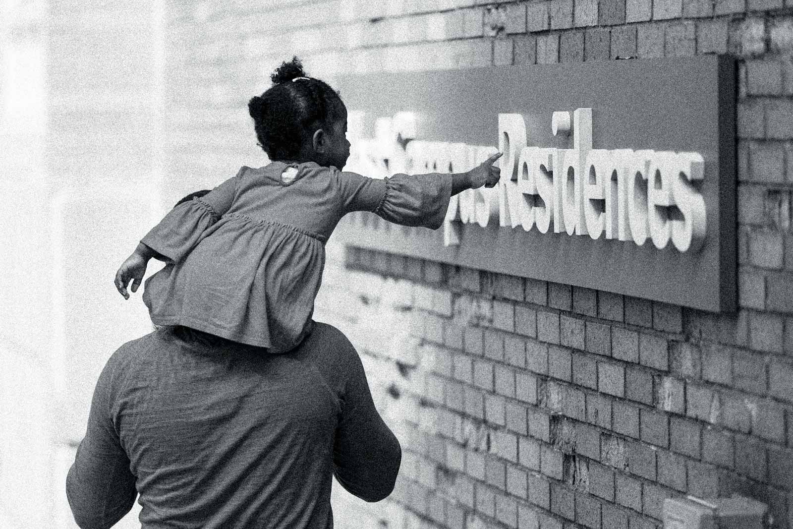 A Black little girl on an adult's shoulder's leans over to point at a BU residence building sign