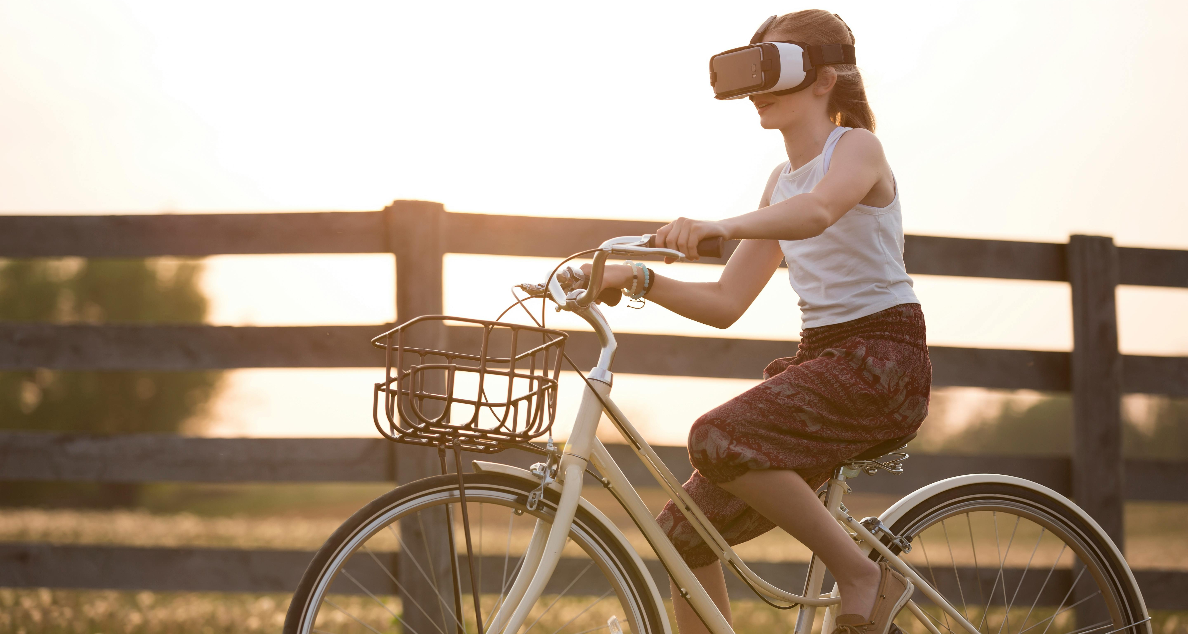 Girl wearing virtual reality goggles while riding a bike in front of a wooden fence