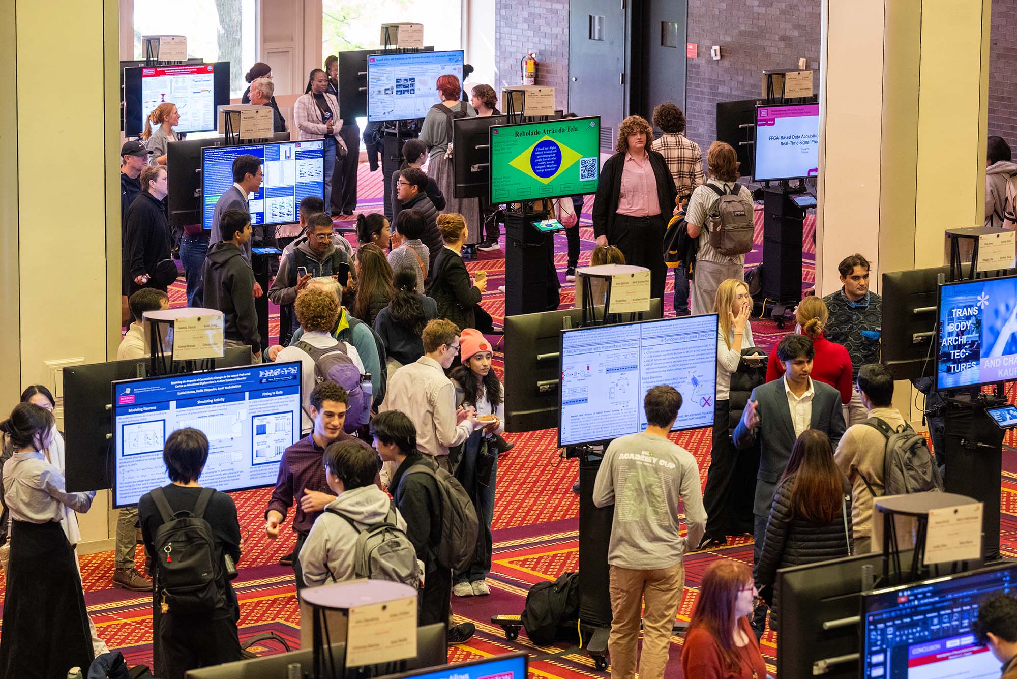 Wide image of BU's UROP Symposium, with many people in a ballroom presenting and looking at poster sessions on digital boards