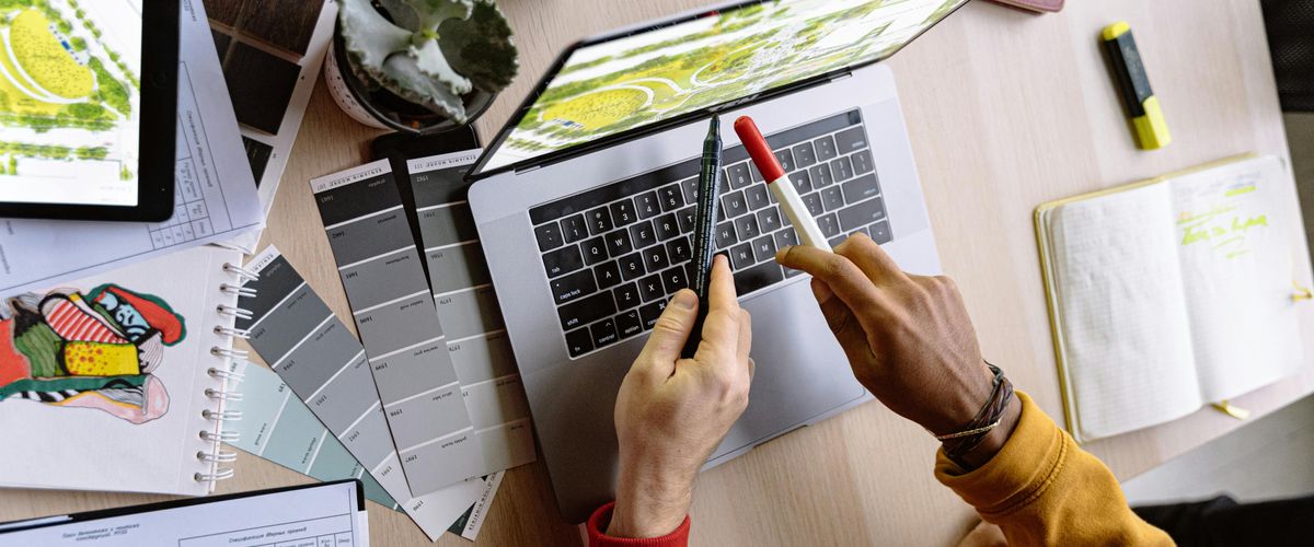 Two hands of different people using pens to point at a laptop surrounded by design supplies
