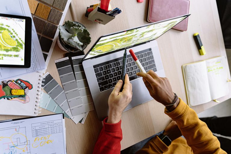 Two hands of different people using pens to point at a laptop surrounded by design supplies