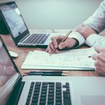 Hands of two people working on budget paperwork with two laptops on a wood desk