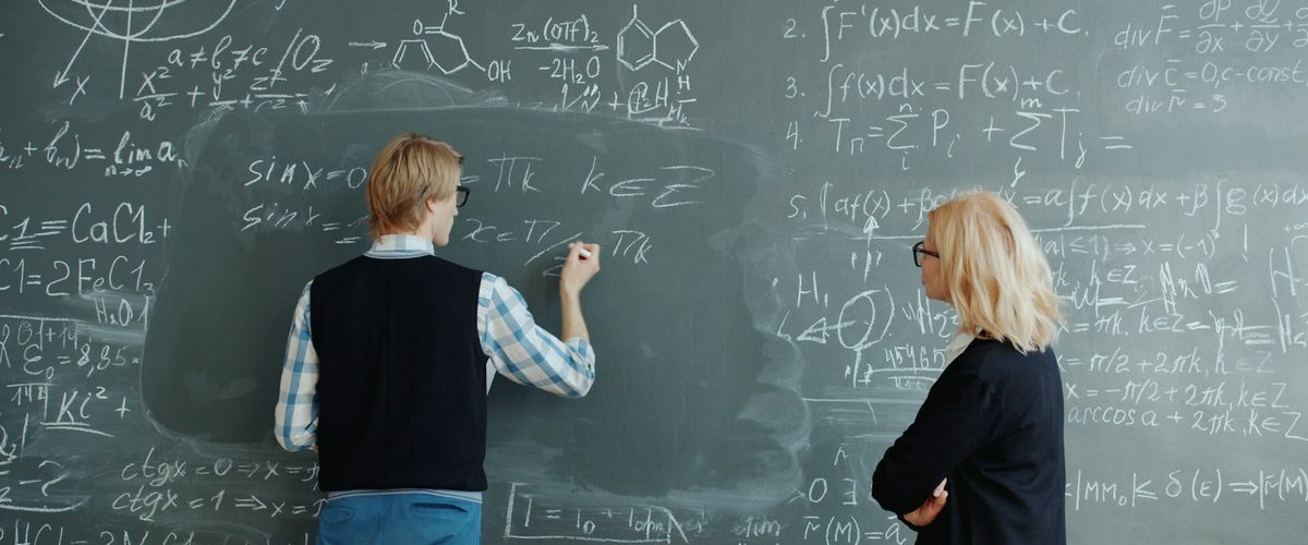 Young male teacher and middle-aged female teacher do calculations on green board with chalk