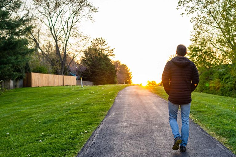 Man walks along an asphalt path through green grass with his back to the camera during golden hour