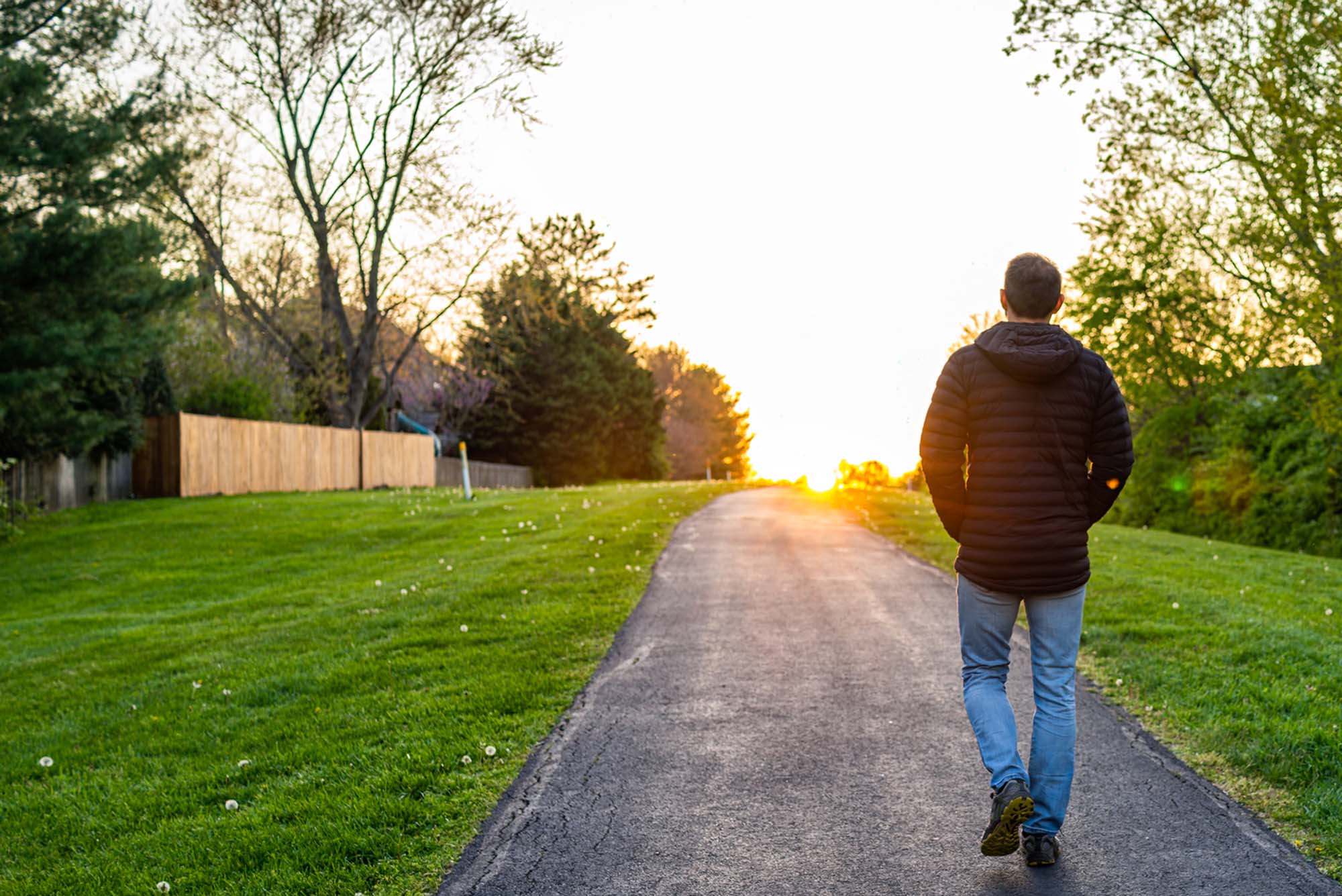 Man walks along an asphalt path through green grass with his back to the camera during golden hour