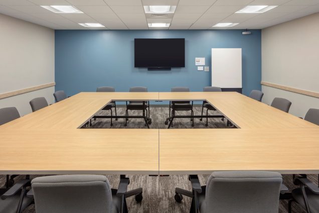 Conference tables setup in boardroom style (square with empty square in the center), a TV mounted on a blue wall, and a marker board in the corner.