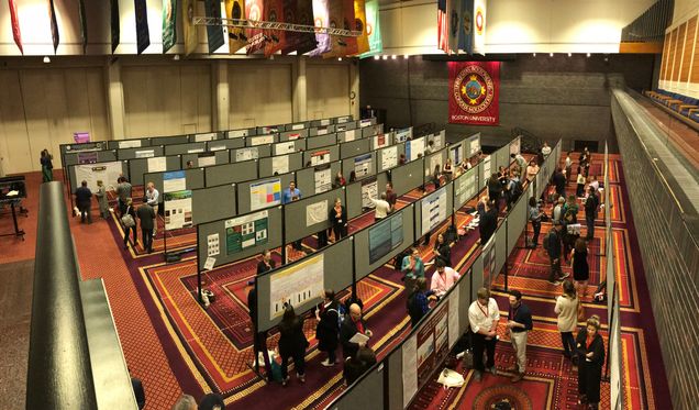 Rows of grey fabric bulletin boards setup for a Poster Session as people walkthrough rows, stopping to discuss posters with researchers.