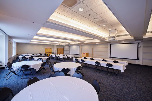 Angular view of Colloquium Room setup with round & rectangular tables with a BU podium and 2 screens for projection.