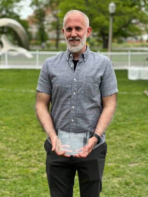 Andrew Vigue poses with curved glass award on the grassy lawn of the BU Beach.