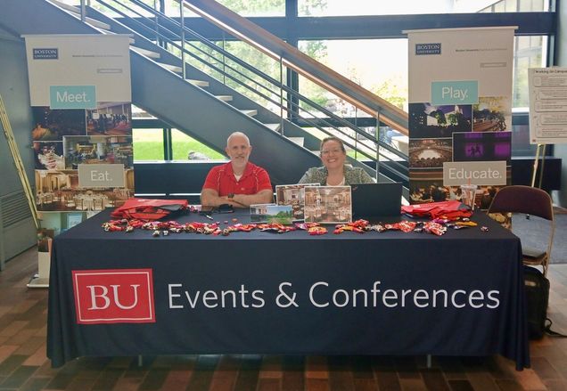 Two staff sitting behind table with black tablecloth that says "BU Events & Conferences"