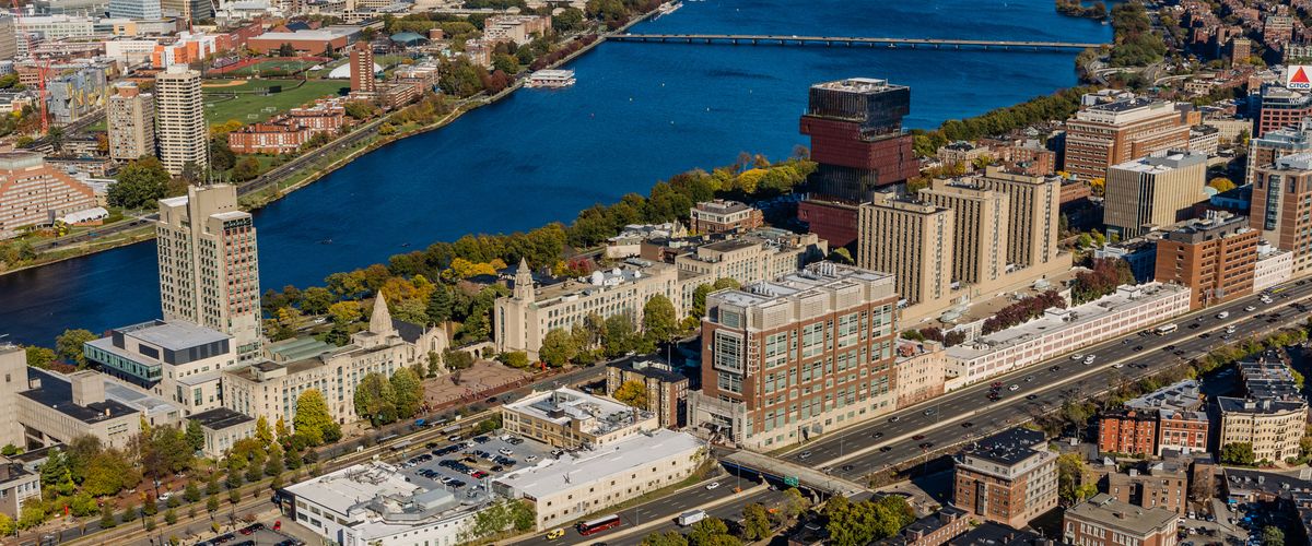 An aerial photograph of the Boston University campus and surrounding areas. The Charles River runs through the center of the image, bordered by university buildings, green spaces, and city infrastructure. Prominent structures and landmarks are visible, with downtown Boston and the skyline in the background.