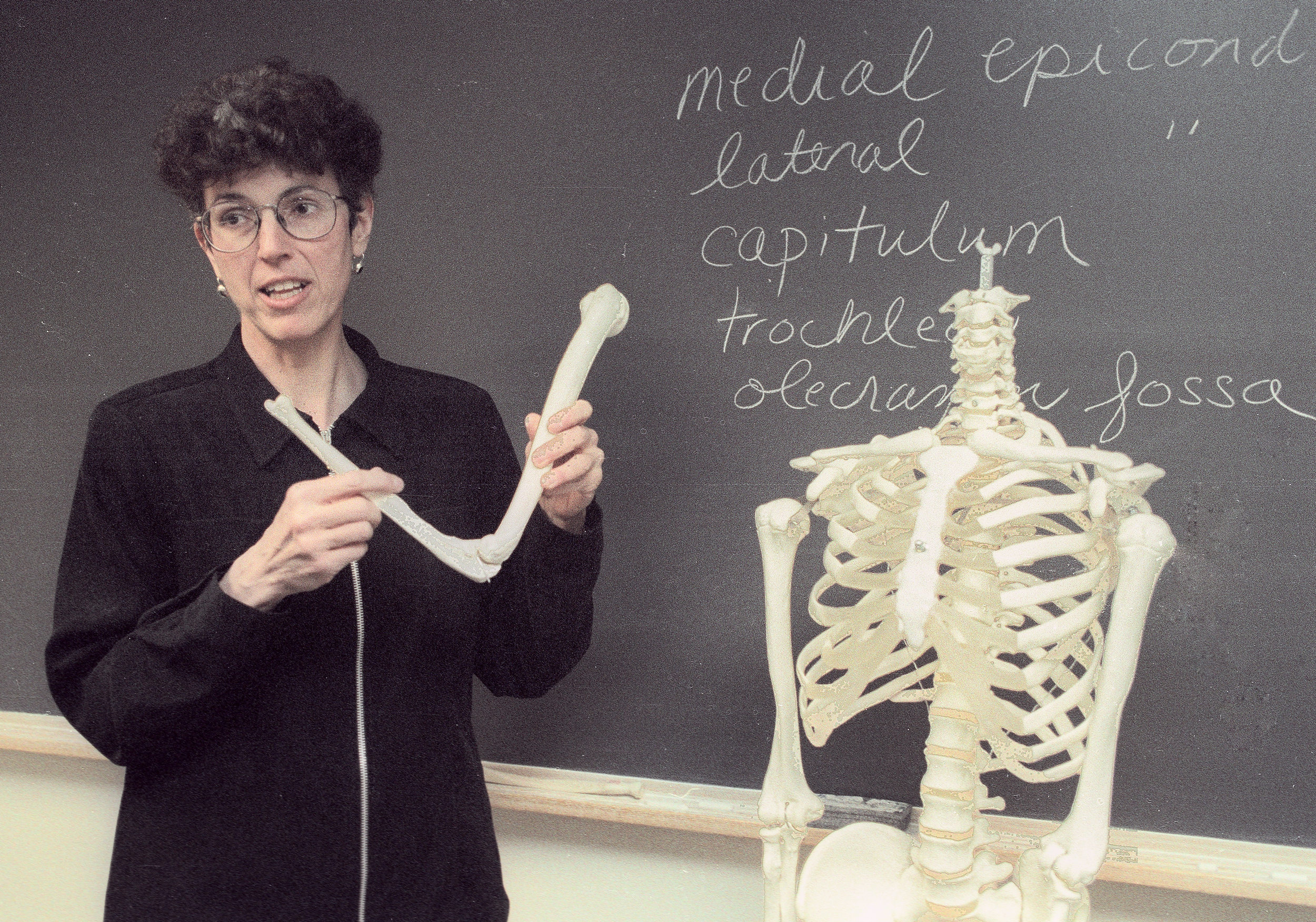 Sargent classroom scene. Professor holding a part of a skeleton with a blackboard behind her.