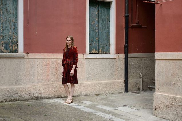 A figure with long, light hair wearing a red velvet dress and low-tan heels walks through a narrow street or courtyard. The courtyard has a tan floor, rose walls to elbow height, and darker pink walls above, interspersed with green shutters, their paint peeling.