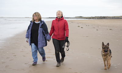 Mature female couple laughing and holding hands walking along the beach on a cold day