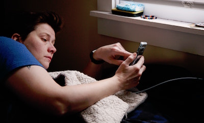 Shane, an immunocompromised nonbinary person, checks their phone in bed while isolating during the COVID-19 pandemic