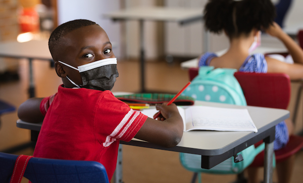 Portrait of african american boy wearing face mask sitting on his desk in class at elementary school