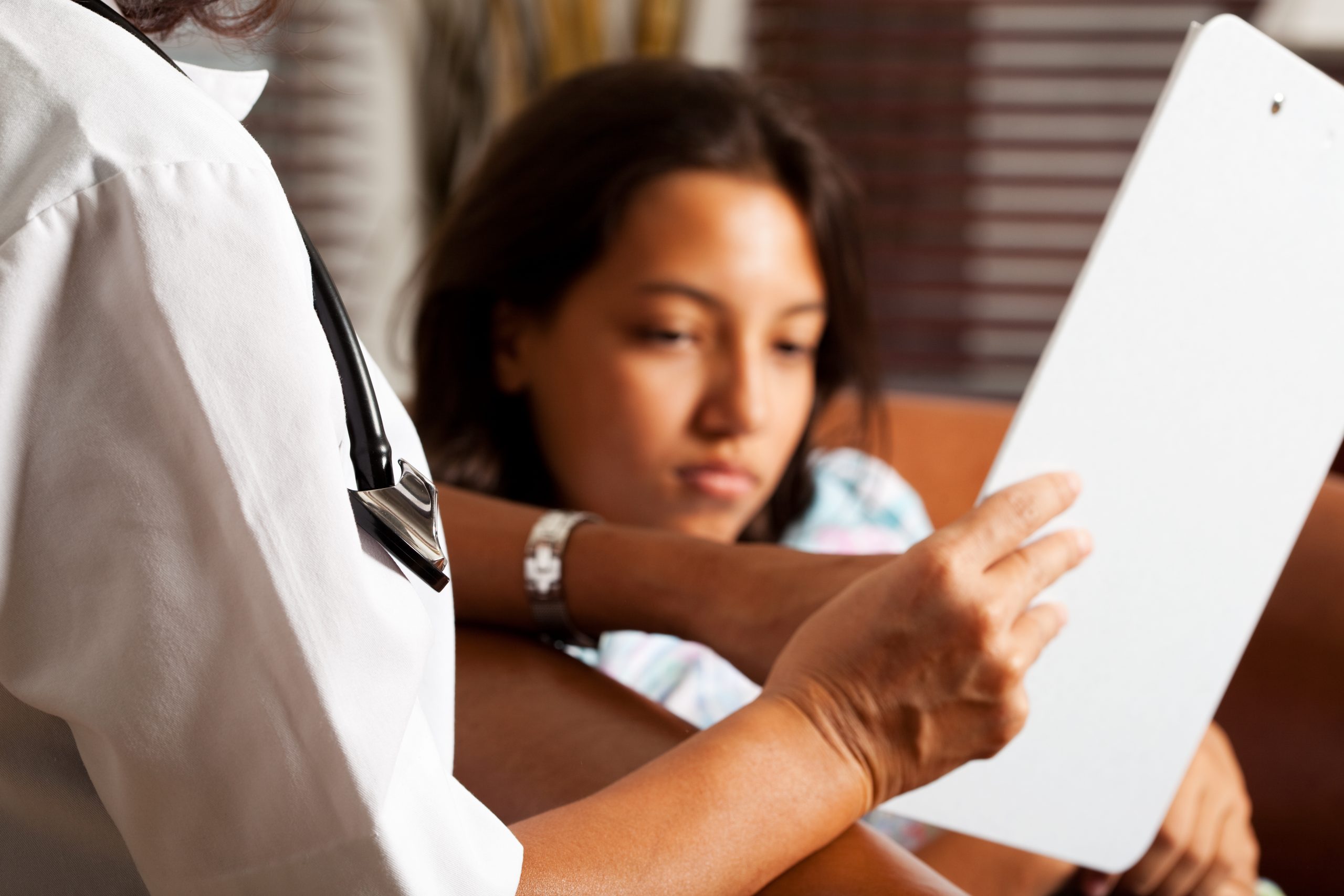 Doctor holding a clipboard, showing it to her child patient and explaining something. Focus on the front (stethoscope and doctor's uniform), patient out of focus in the background