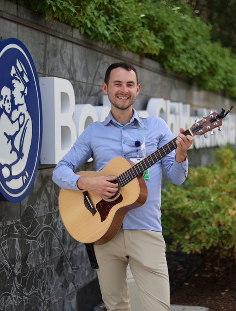 Mark Fuller smiles holding a guitar in from of the Boston Children’s Hospital