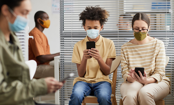 Front view portrait of multi-ethnic group of people using smartphones while waiting in line at medical clinic, all wearing masks