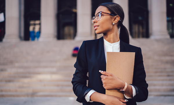 Female lawyer in stylish formal suit, holding folder, looking away standing against courthouse