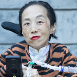 Photo of Alice Wong, an Asian American disabled woman in a power chair. She is wearing an orange and black tiger-striped sweater, black pants, a bold red lip color and a trach at her neck. In the background is a gray cement wall with greenery. Photo credit: Eddie Hernandez Photography.