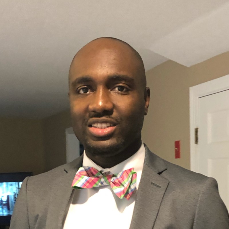 Jean-Claude Regis, wearing a pink and green bowtie, a white collared shirt, and grey suit jacket, smiles at the camera