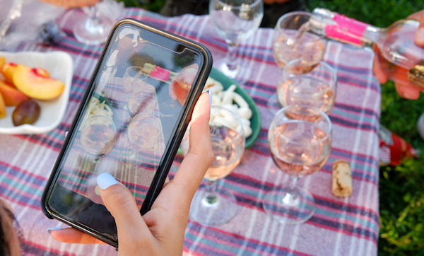 Woman's hand taking a photo of glasses of rose wine and cheese board with her smartphone on a picnic table