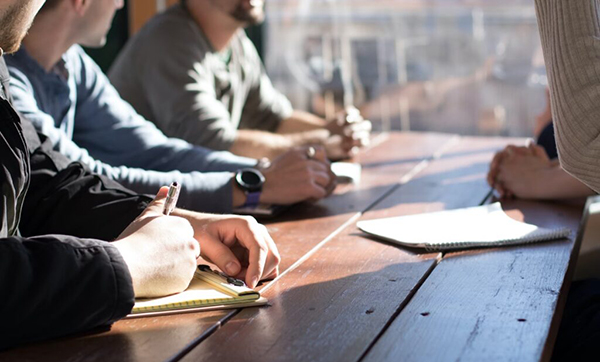 Four people sit at a wooden table with notepads