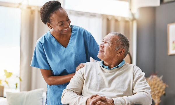 Elderly man sits in a chair and smiles up at nurse with her hand on his shoulder