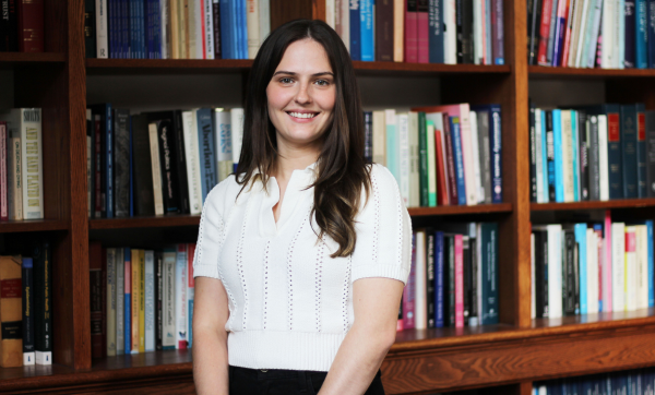 Dana Vasers, wearing a white, short-sleeved sweater, stands in front of a large bookcase and smiles at the camera