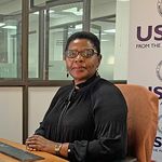 Muka Chikuba-McLeod sits at a desk in front of a USAID sign