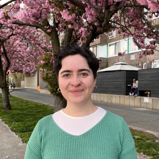 Maria Bermudez, with short, dark hair, wearing a green sweater and white shirt, stands in front of a flowering cherry blossom tree