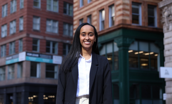 Leedya Senbetta smiles at the camera while standing on a city block wearing a black blazer and white collared shirt.