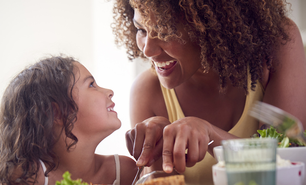 Mother Helping Daughter To Cut Food Sitting Around Table At Home Eating Family Meal Together