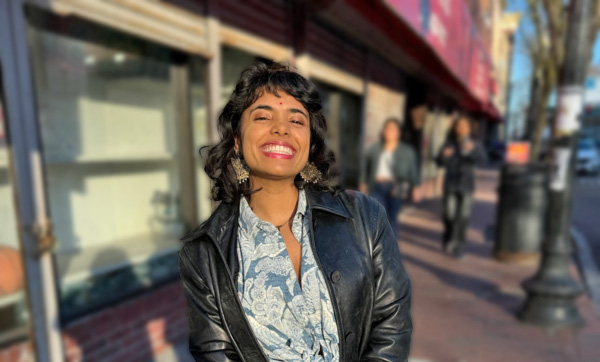Aisha Ghorashian stands on a sidewalk smiling at the camera, she is wearing a leather jacket and white and blue blouse