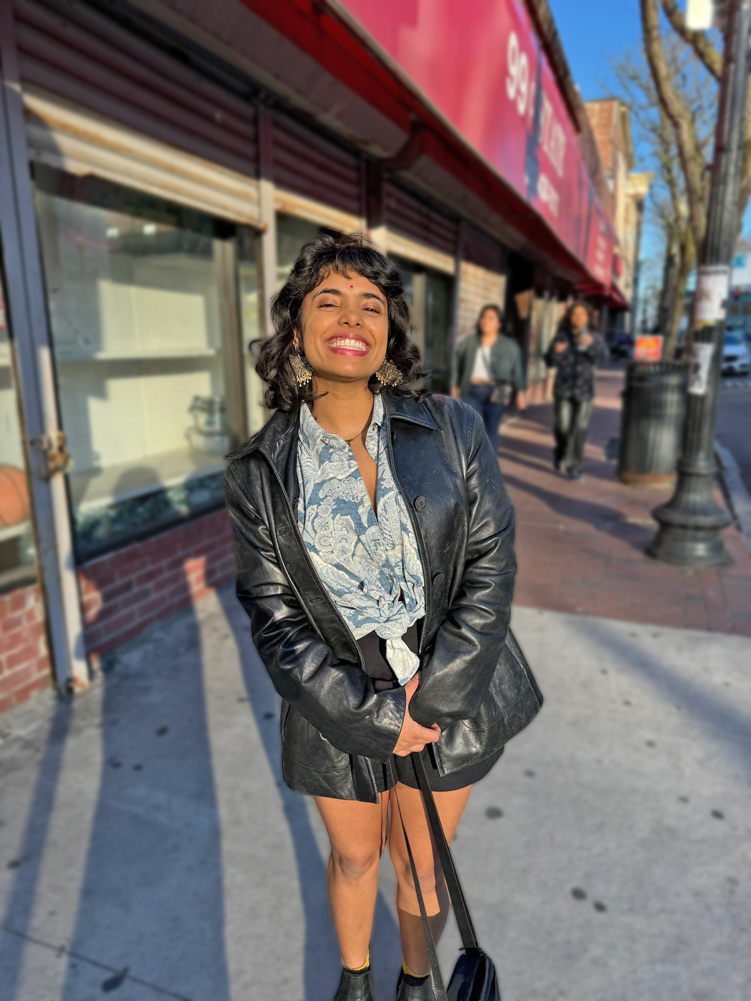 Aisha Ghorashian stands on a sidewalk smiling at the camera, she is wearing a leather jacket and white and blue blouse