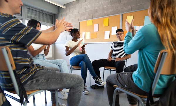 Latino male teen student receives praise from classmates. Multiracial group of high school students sitting in a circle clapping together celebrating. Togetherness.