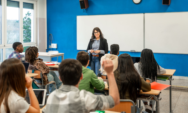 Multi-ethnic group of teenage students listening to their teacher's lecture and raising hands to answer questions in high school classroom
