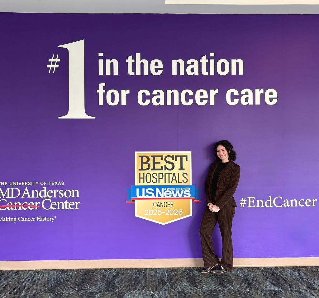 Carla Irizarry-Delgado, dressed in a brown suit, stands in front of a purple wall that reads "The University of Texas MD Anderson Cancer Center," "#1 in the nation for cancer care," and "#EndCancer" with the US News logo for best hospitals for cancer.