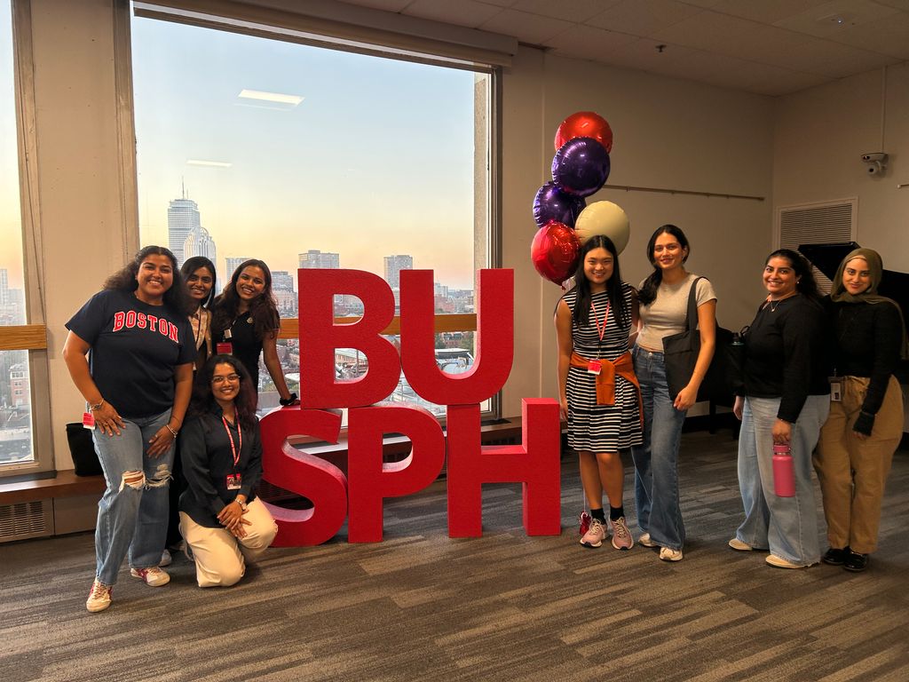 A group of students pose with the letters "BUSPH" and a bundle of balloons. The sun sets on Boston through the window in the background.