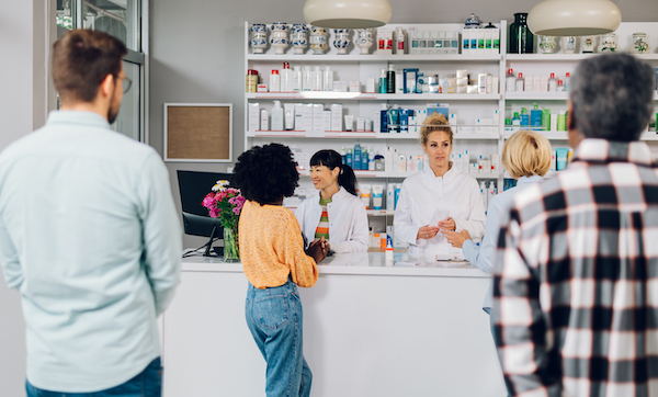 A queue of people standing in a pharmacy while shopping for medicine, drugs, vitamins, supplements, health care beauty products. Focus on a two woman pharmacist working while standing at the counter.