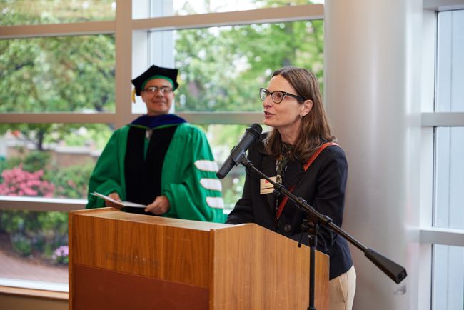 Sonia Mee, assistant director of online programs, speaks at a podium. Dean Delva stands behind her, smiling.