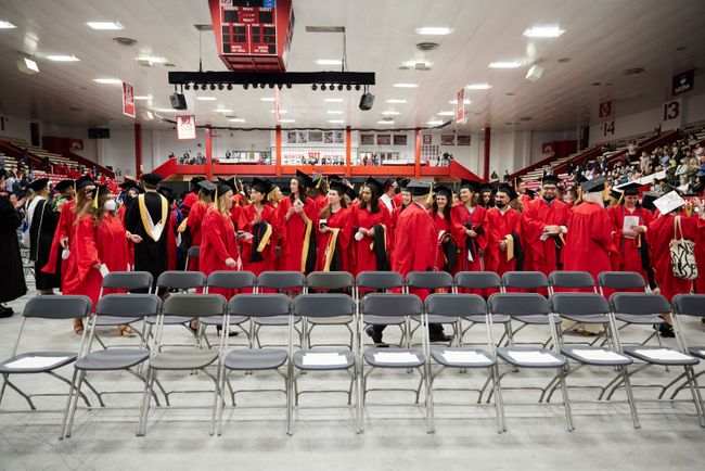 Grads stand at their seats as the procession continues. Two rows of empty chairs are in the foreground, waiting for faculty to return from the procession