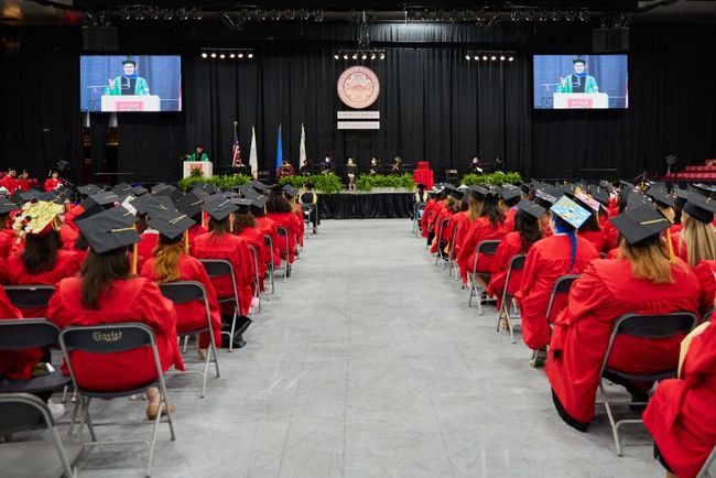 Photo looks down the center aisle at commencement. Grads are seated on both sides and Dean Delva is at the podium and shown on two jumbotron screens.