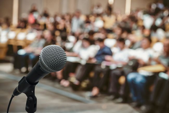 Photo shows microphone in foregrounnd with blurred conference hall audience in background